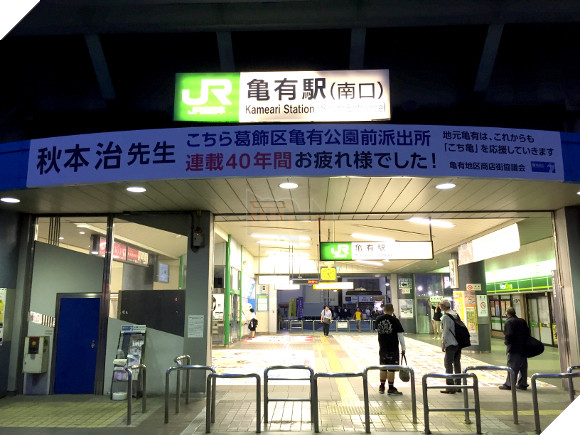 
Một tấm biểu ngữ trắng lớn được đặt ở ngay trước cửa ra vào nhà ga Kameari với lời cảm ơn và chúc mừng tác giả Akimoto Osamu vì 40 năm làm việc cống hiến hoàn thành bộ manga
